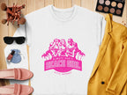 A Garment Graphics white sweatshirt with a pink graphic of three women and palm trees, labeled Always Party Beach Girl, sits alongside makeup brushes, eyeshadow, lipstick, sunglasses, a yellow jacket, pink shoes, and a compact mirror on a wooden surface.
