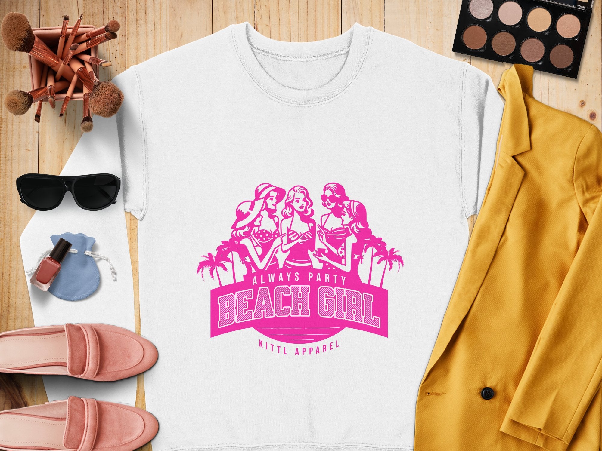 A Garment Graphics white sweatshirt with a pink graphic of three women and palm trees, labeled Always Party Beach Girl, sits alongside makeup brushes, eyeshadow, lipstick, sunglasses, a yellow jacket, pink shoes, and a compact mirror on a wooden surface.