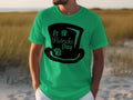 A person sports a green t-shirt by Garment Graphics featuring a black top hat with St Patricks Day text and two small shamrocks, perfect for celebrating the festive spirit on grass or sand while searching for a four-leaf clover.
