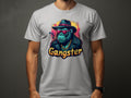 A person is sporting a white Garment Graphics Gangster Gorilla T-Shirt with urban style. This classic fit tee displays a gorilla dressed as a gangster—complete with hat, sunglasses, coat, gold chain, and smoking cigar. The word Gangster stands boldly beneath the illustration.