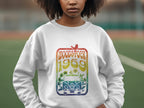 An individual stands outdoors in a white Garment Graphics sweatshirt with retro vibes, showcasing a colorful design that includes Woodstock 1969 above an illustrated van and flowers.
