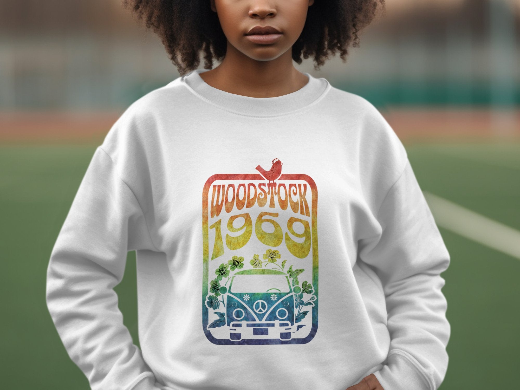 An individual stands outdoors in a white Garment Graphics sweatshirt with retro vibes, showcasing a colorful design that includes Woodstock 1969 above an illustrated van and flowers.