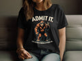 Indoors, a person with long, wavy hair wears a Garment Graphics black T-shirt displaying the Admit It Bigfoot design. The playful image shows Bigfoot giving a peace sign with the text ADMIT IT. LIFE WOULD BE BORING WITHOUT ME.