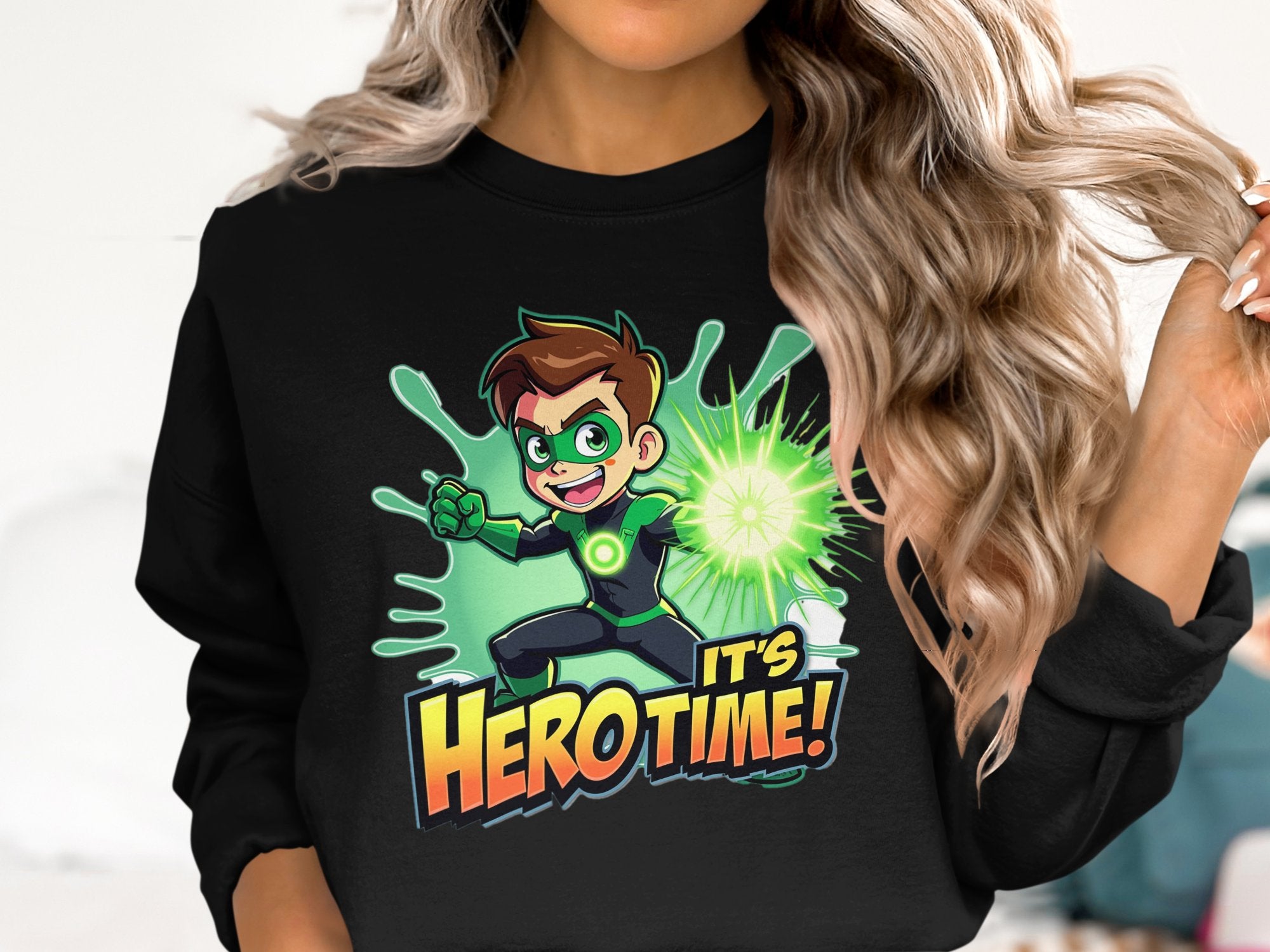 A person in a Garment Graphics black sweatshirt features a cartoon superhero with brown hair, green mask, and green energy powers. Bold Its Hero Time! text in orange and yellow complements their wavy blonde hair. Unisex sizing is ideal for them.