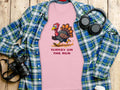 The Garment Graphics classic fit pink T-shirt features a humorous TURKEY ON THE RUN design, displayed on wood with a green and blue plaid shirt, camera, headphones, and coffee cup.