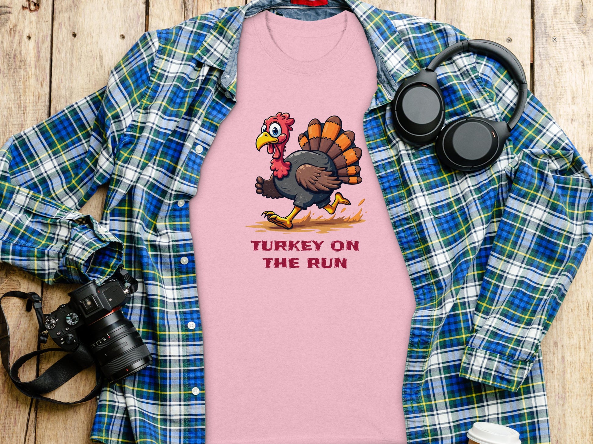 The Garment Graphics classic fit pink T-shirt features a humorous TURKEY ON THE RUN design, displayed on wood with a green and blue plaid shirt, camera, headphones, and coffee cup.