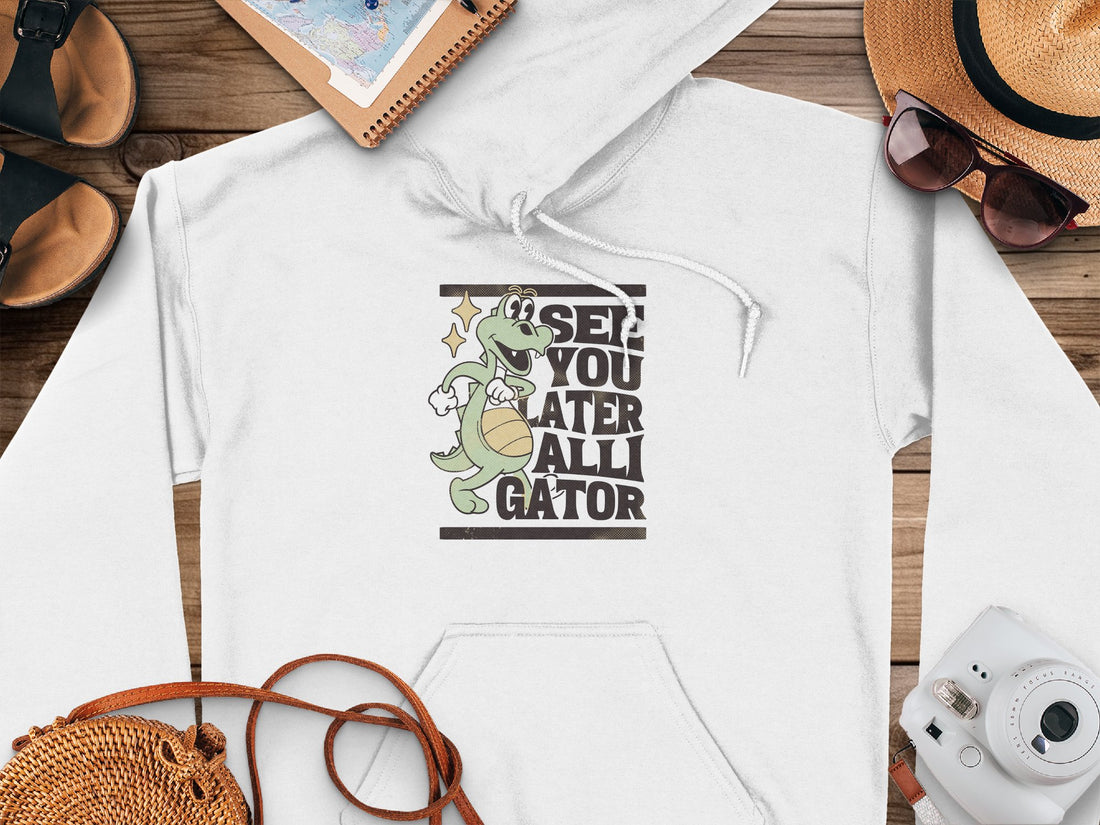 A person wears a classic fit light pink hoodie by Garment Graphics, featuring a gator graphic and the text SEE YOU LATER ALLIGATOR, sitting against a festive checkered background, exuding comfort and style.