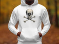 A person stands in a blurred outdoor setting with vibrant autumn foliage, wearing a fashion-forward white hoodie by Garment Graphics featuring a skull and crossbones design, their hands resting casually in the hoodies front pocket.