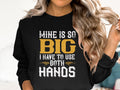 A person with long, wavy blonde hair is wearing a distinctive sweatshirt from Garment Graphics with the bold statement: MINE IS SO BIG I HAVE TO USE BOTH HANDS, embellished with decorative accents.