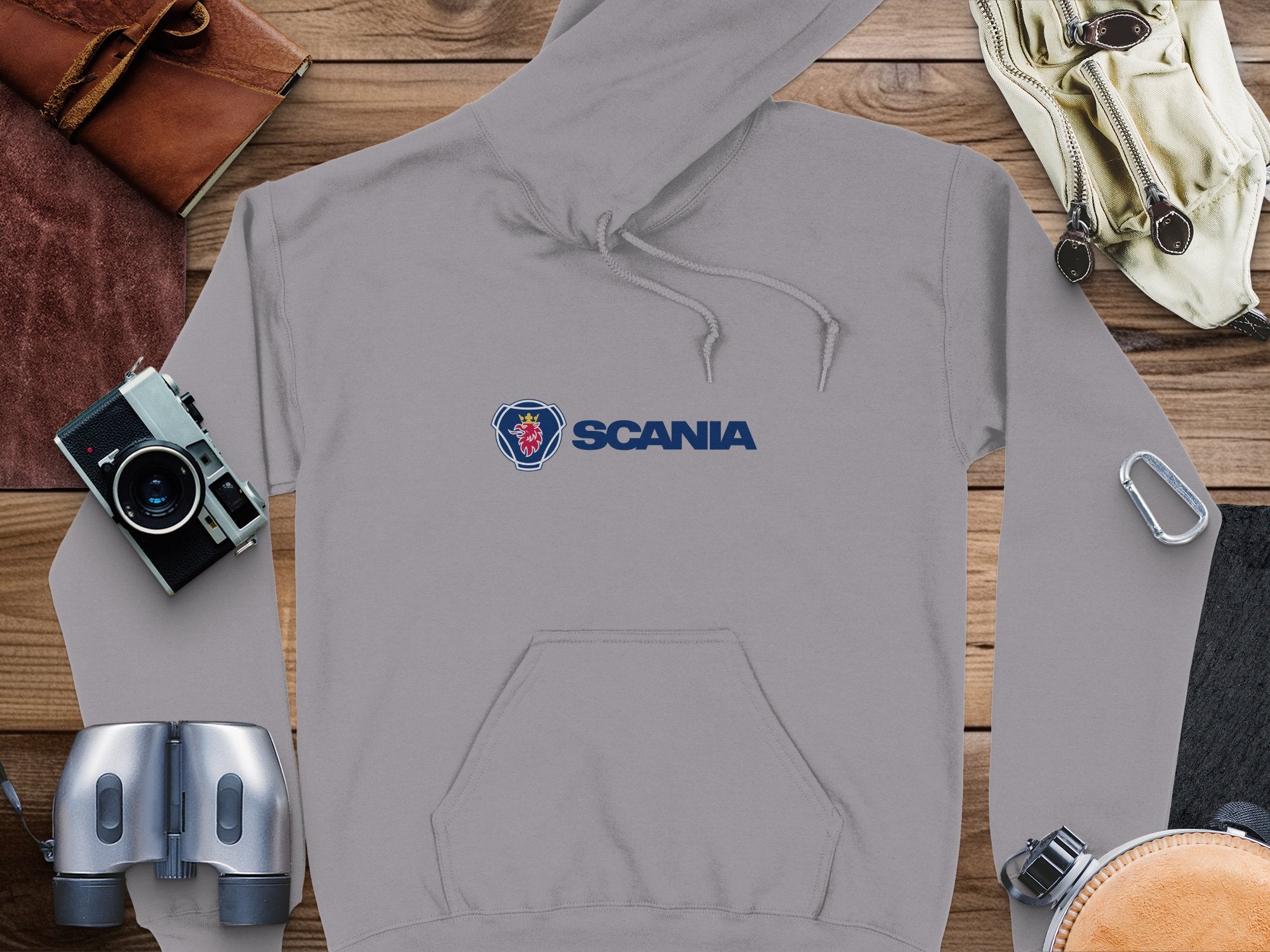 A stylish gray hoodie from Garment Graphics with the Scania logo featuring a griffin head is laid on wood, surrounded by a camera, binoculars, gloves, carabiner, leather wallet, and folded map—ideal for fans of branded hoodies.