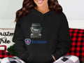 A brown-haired woman smiles while sitting, wearing a black Garment Graphics Scania truck hoodie with a graphic design. A red-and-black checkered pillow and a Christmas-themed item create the perfect scene for truck enthusiasts in the background.