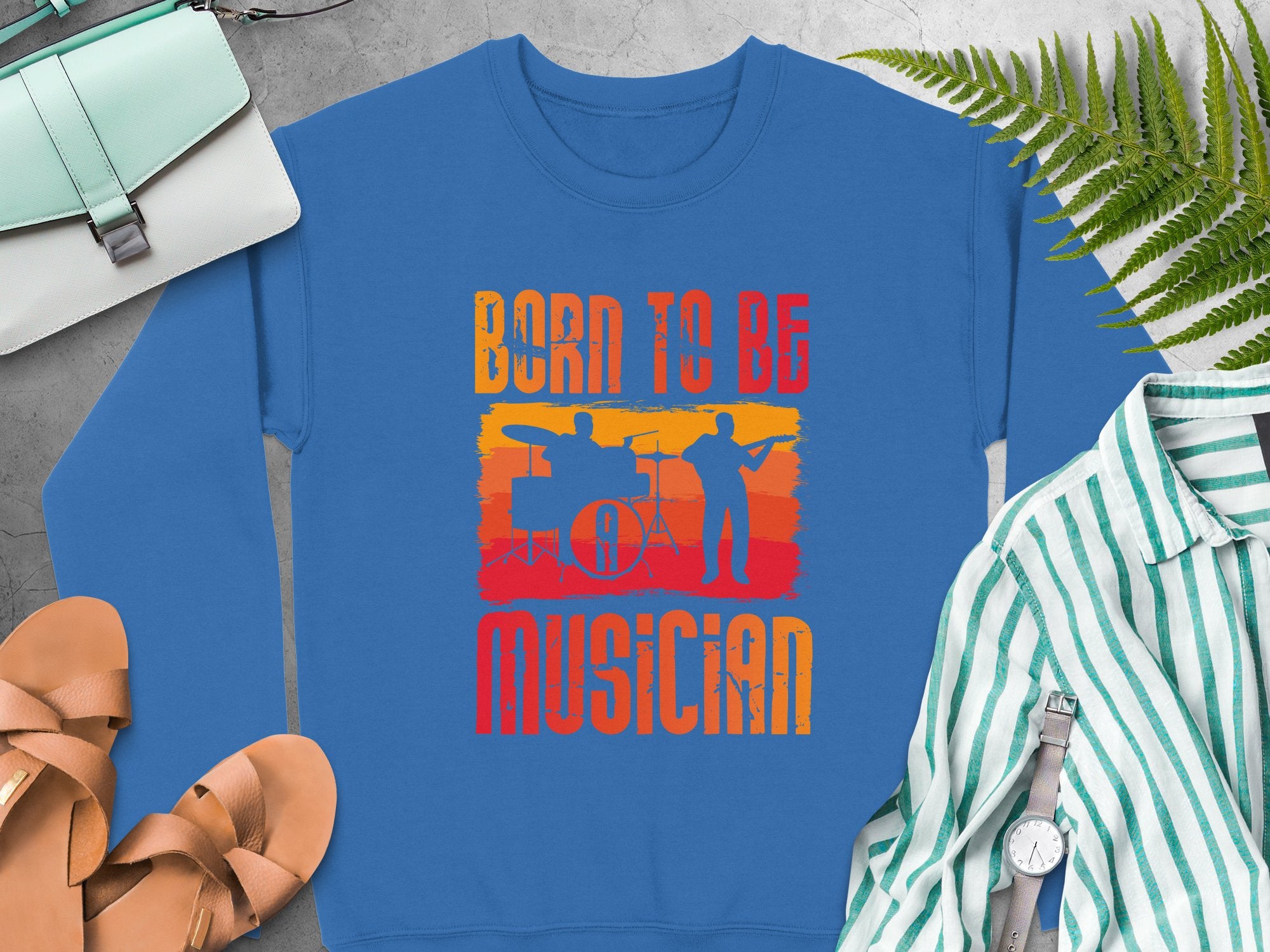 A blue unisex sweatshirt from Garment Graphics sits beside sandals, a watch, a striped shirt, a fern, and a handbag. Ideal for music enthusiasts, it showcases Born to Be Musician with silhouettes of a drummer and guitarist against a sunset backdrop.