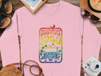 Garment Graphics pink sweatshirt showcases a Woodstock 1969 design with a rainbow van and bird. Its creatively styled on wood with a hat, sunglasses, sandals, camera, and notebook for ultimate retro vibes.