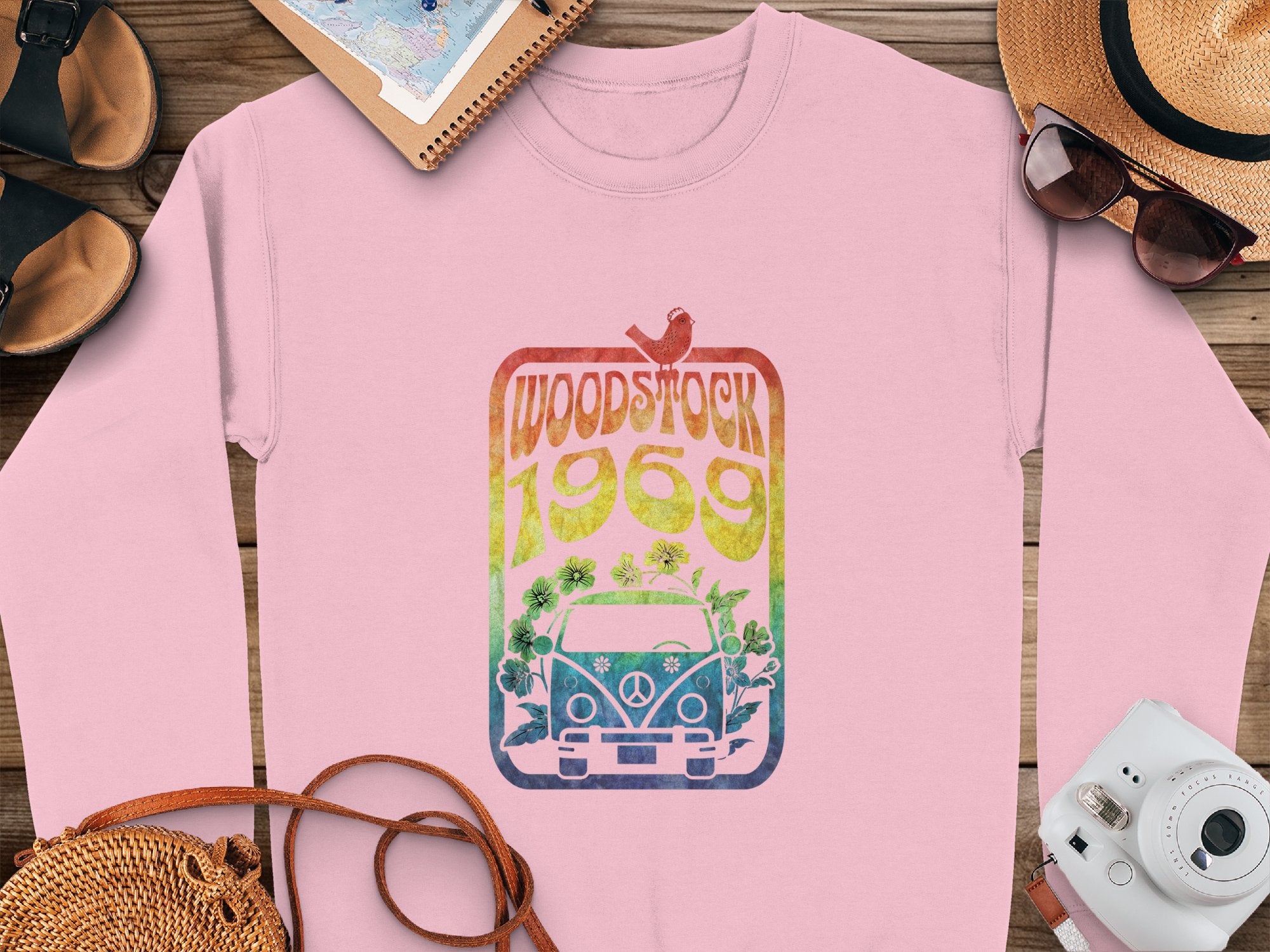 Garment Graphics pink sweatshirt showcases a Woodstock 1969 design with a rainbow van and bird. Its creatively styled on wood with a hat, sunglasses, sandals, camera, and notebook for ultimate retro vibes.