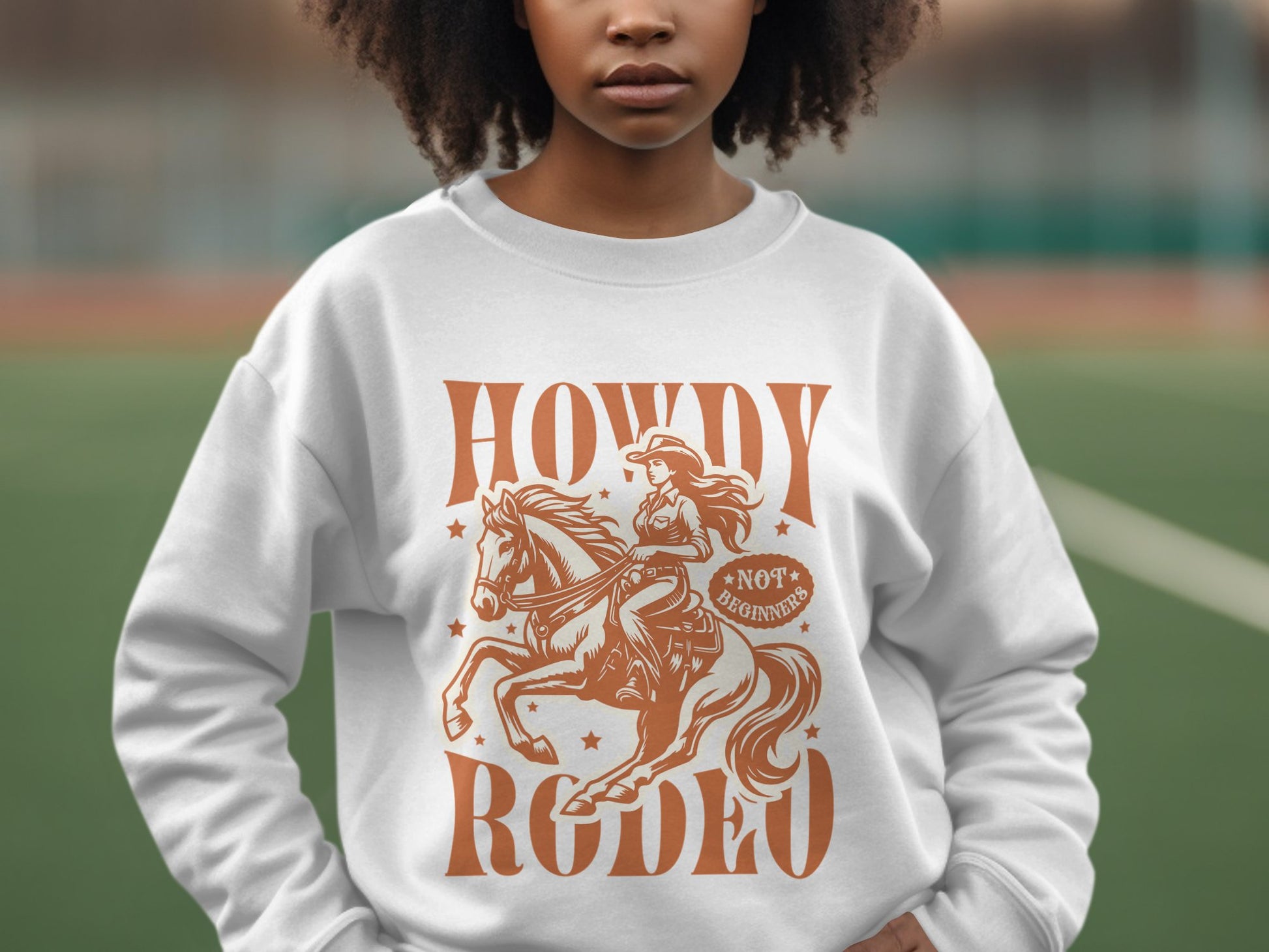 A person outdoors wears a white Garment Graphics sweatshirt with a brown cowgirl and horse graphic, surrounded by stars and featuring the words Howdy Rodeo and Not for Beginners. Perfect for rodeo enthusiasts looking to embrace the cowboy spirit in style.