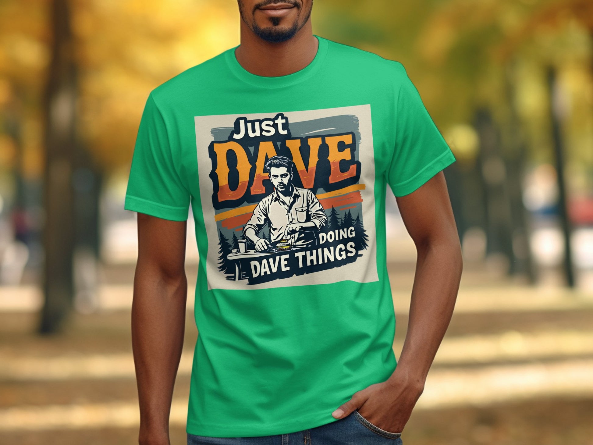 A person wears a classic fit green graphic T-shirt by Garment Graphics, featuring the text Just Dave Doing Dave Things, set against a backdrop of blurred autumn trees.