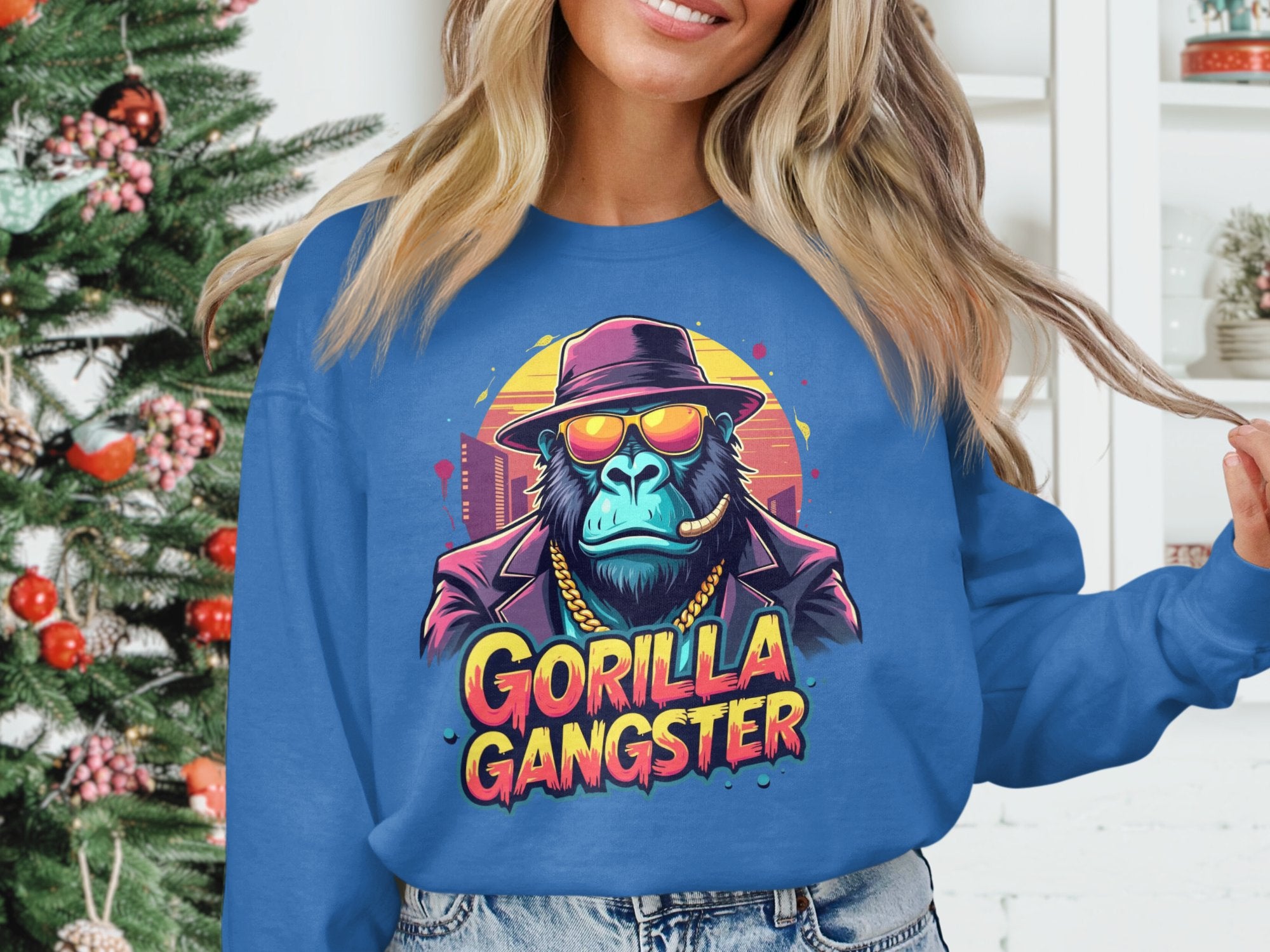 Wearing a Garment Graphics blue sweatshirt with a Gorilla Gangster in hat, sunglasses, and gold chain, someone stands by a festively decorated Christmas tree, showcasing an urban style.
