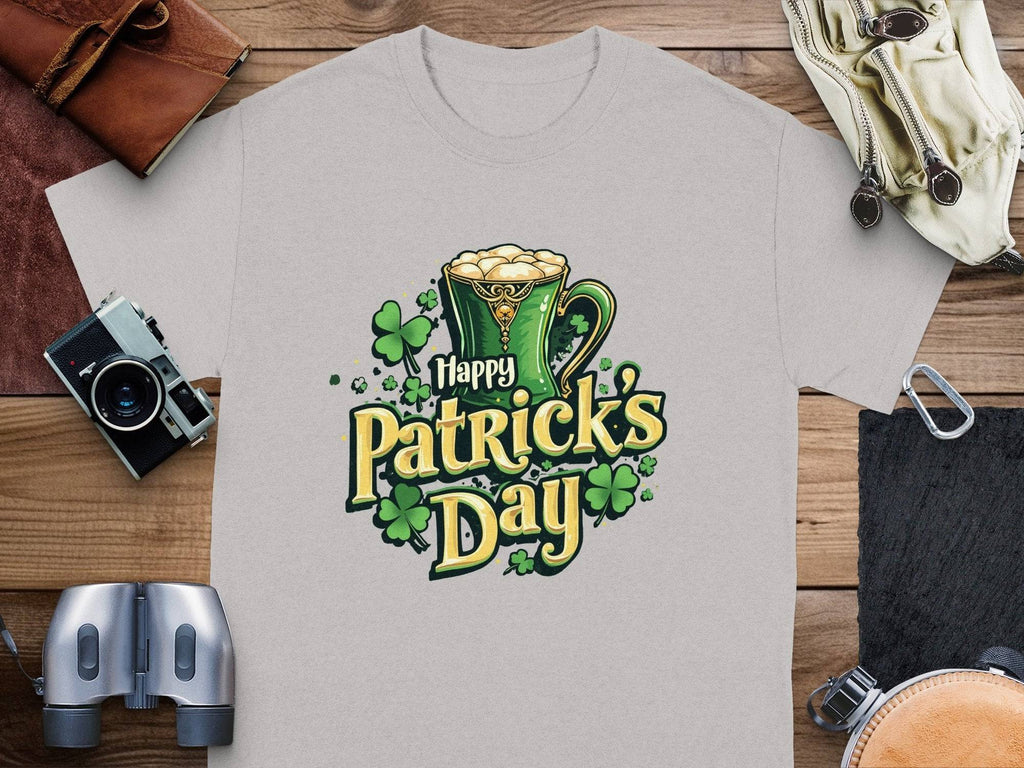 The Garment Graphics gray T-shirt has Happy Patricks Day in green and yellow, highlighted by a mug of beer and shamrocks. Ideal for celebrations, it rests on a wooden surface with a camera, binoculars, gloves, and a leather pouch.