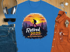 A flat lay features Garment Graphics blue T-shirt with a hiking silhouette and Retired 2025, Not My Problem Anymore text, celebrating retirement. Its paired with headphones, sunglasses, jeans, keys, a watch, and an orange shirt on a wood surface.