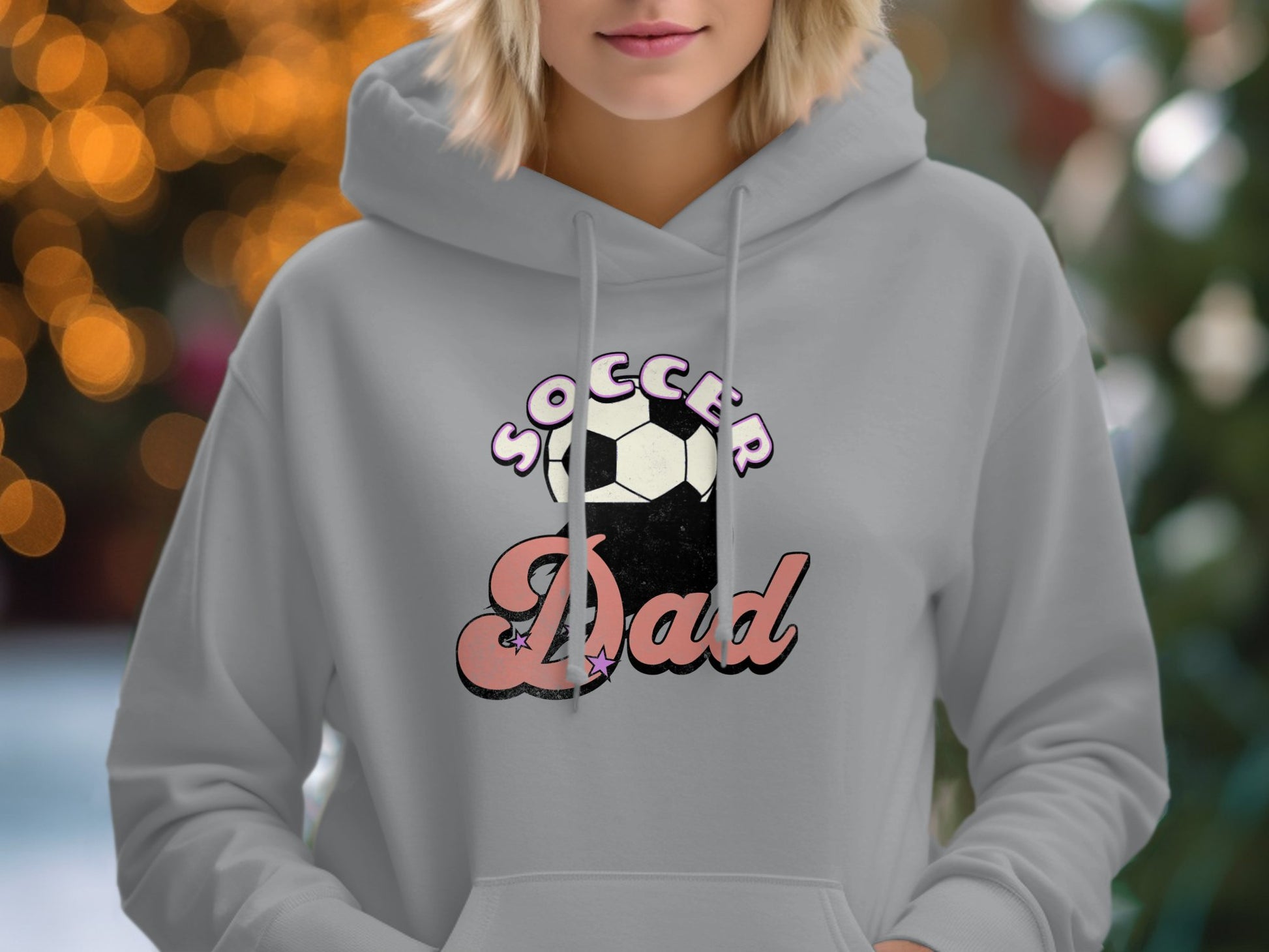 A person in a light gray Garment Graphics hoodie featuring Soccer Dad next to a soccer ball graphic. The softly blurred bokeh-light background adds warmth and charm to this stylish piece, ideal for proud soccer parents.