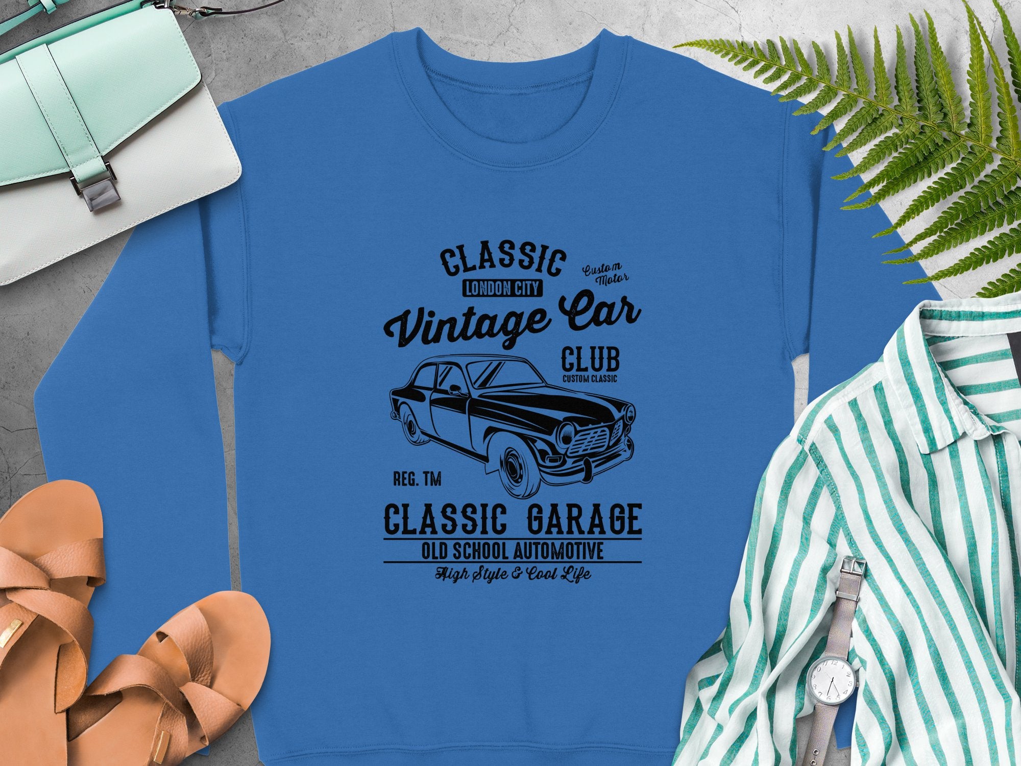 The Garment Graphics blue Vintage Car Sweatshirt features a Classic Vintage Car Club graphic with a car illustration and text: Classic London City, Classic Garage, and Old School Automotive. Nearby are shoes, a purse, and a striped shirt on the gray surface.