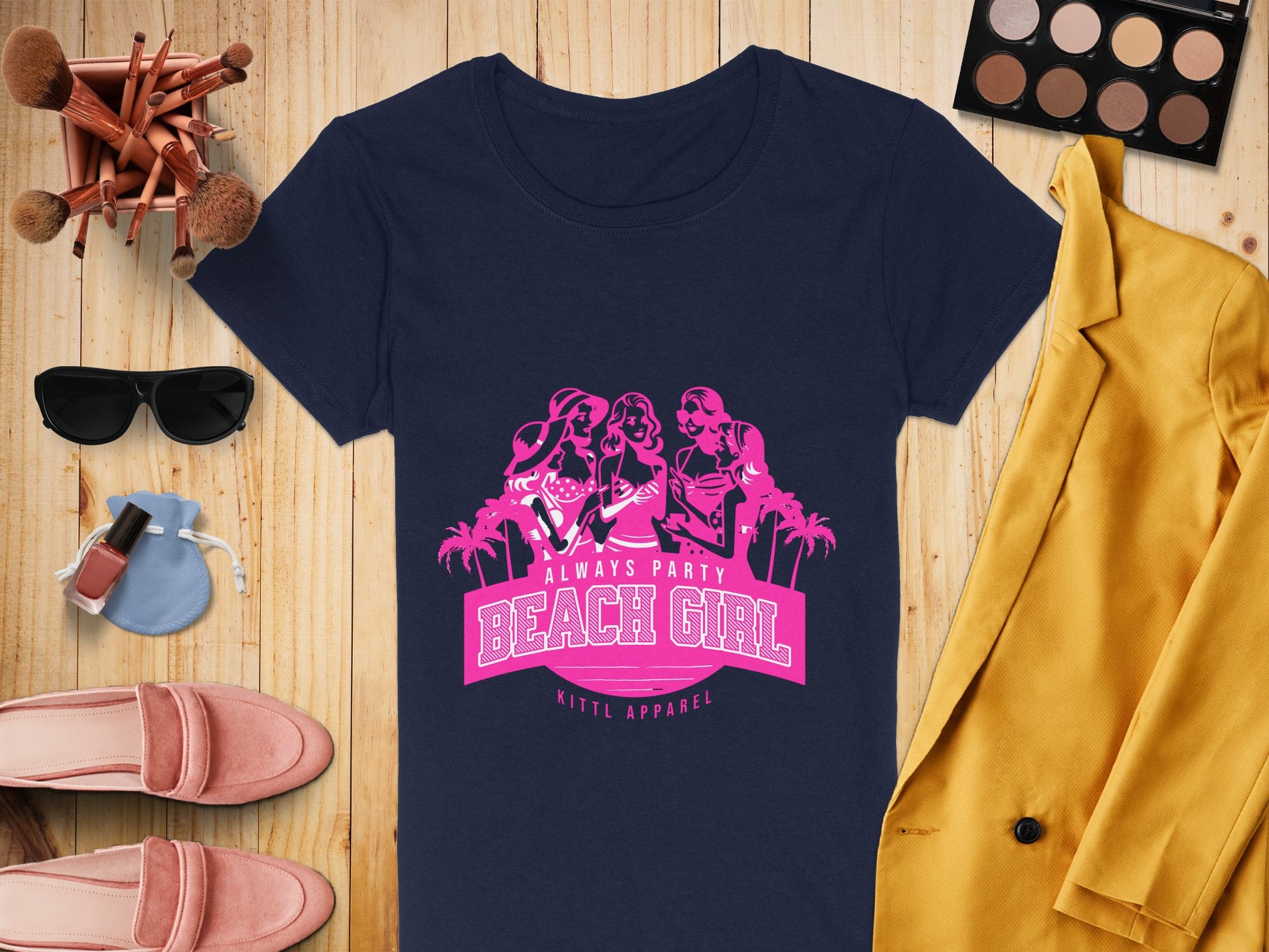Flat lay of a navy Beach Girl T-Shirt by Garment Graphics, with a yellow blazer, next to an eyeshadow palette, makeup brushes, sunglasses, perfume bottle, nail polish, and pink loafers on a wooden surface.