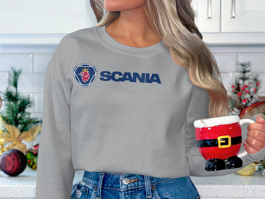 In a festive kitchen, someone holds a Santa-shaped mug, wearing a grey sweatshirt with the Scania logo from Garment Graphics and blue jeans. The unisex sizing offers comfort as Christmas decorations like pine arrangements and ornaments add brightness to the scene.