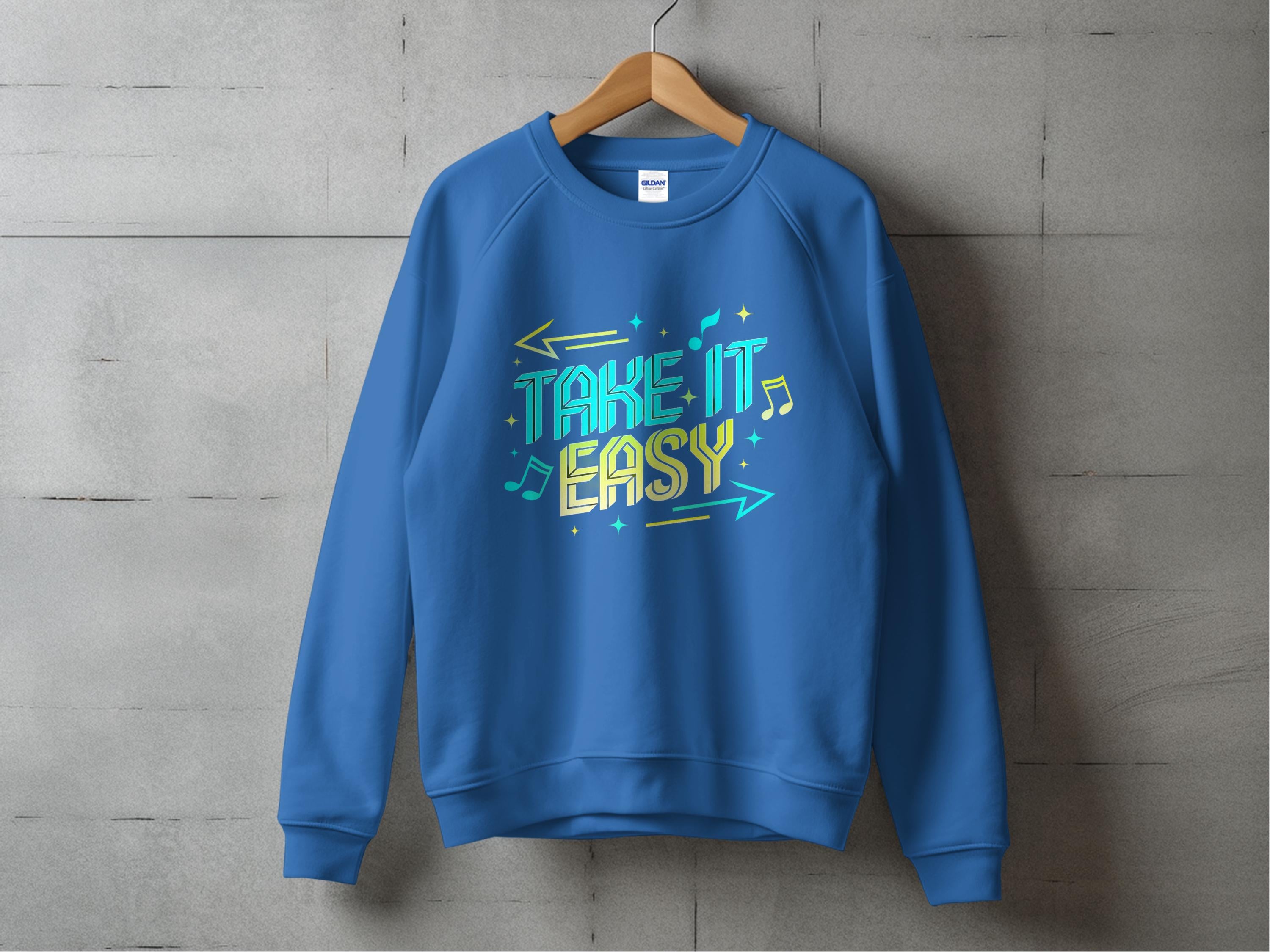 A blue Garment Graphics unisex sweatshirt, featuring a Take It Easy graphic with colorful letters, arrows, and musical notes, hangs on a wooden hanger against a concrete wall—ideal for your casual wear collection.