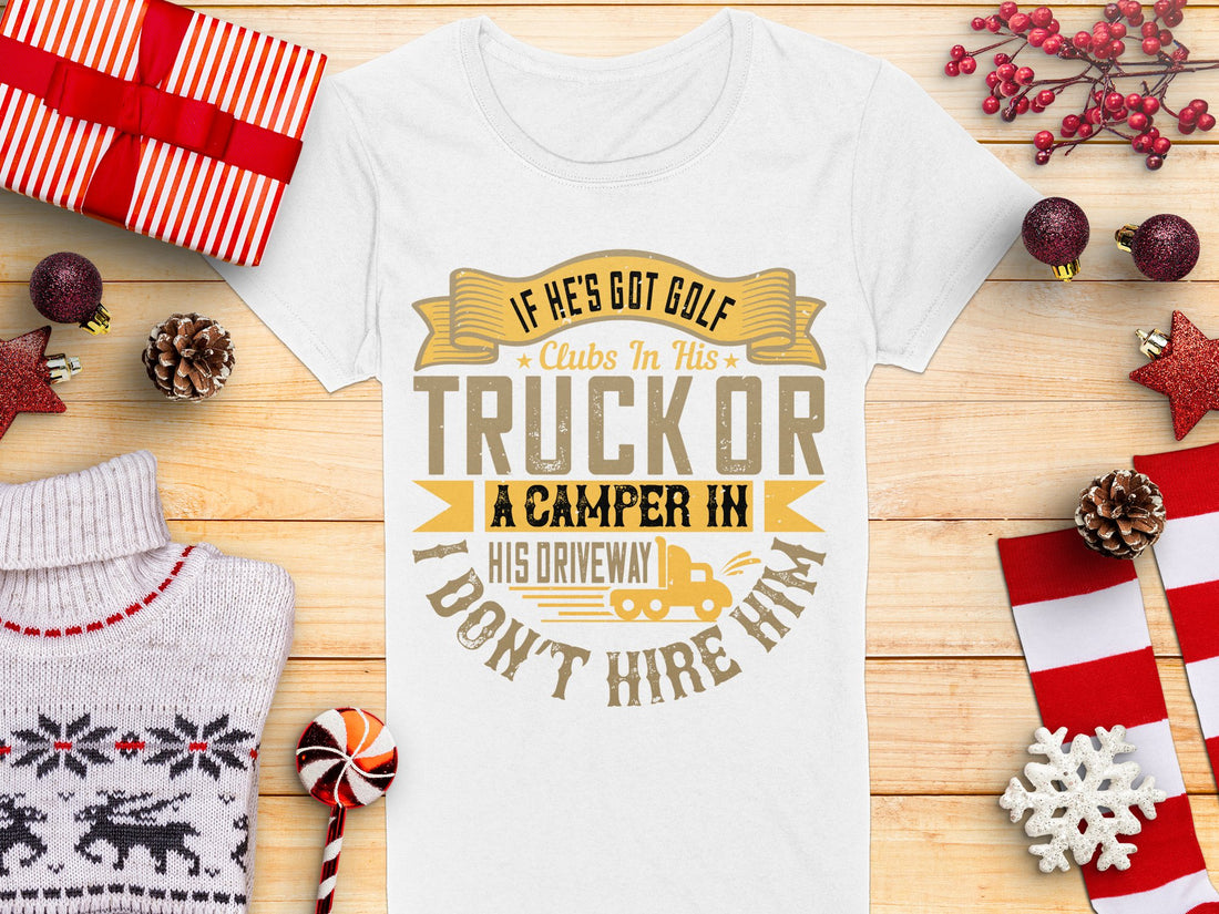 A womens white tee from Garment Graphics displays a humorous golf graphic with the text, If hes got golf clubs in his truck or a camper in his driveway I dont hire him, set against wrapped gifts, a knitted sweater, candy cane, and festive decorations on a wooden backdrop.