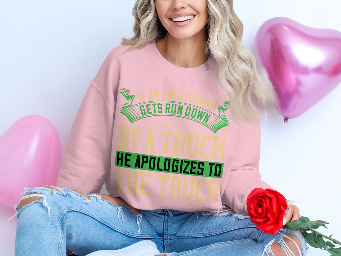 This Garment Graphics unisex sweatshirt flaunts a bold and humorous design with the phrase: If an Englishman gets run down by a truck, he apologizes to the truck, showcased on a wooden surface with sunglasses, makeup items, a yellow jacket, and pink shoes adorning it.