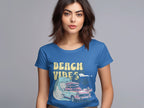 A woman with shoulder-length brown hair is wearing a blue Garment Graphics t-shirt featuring a retro surf design with a surfboard, car, waves, palm trees, and the text Beach Vibes. The background is plain and neutral.