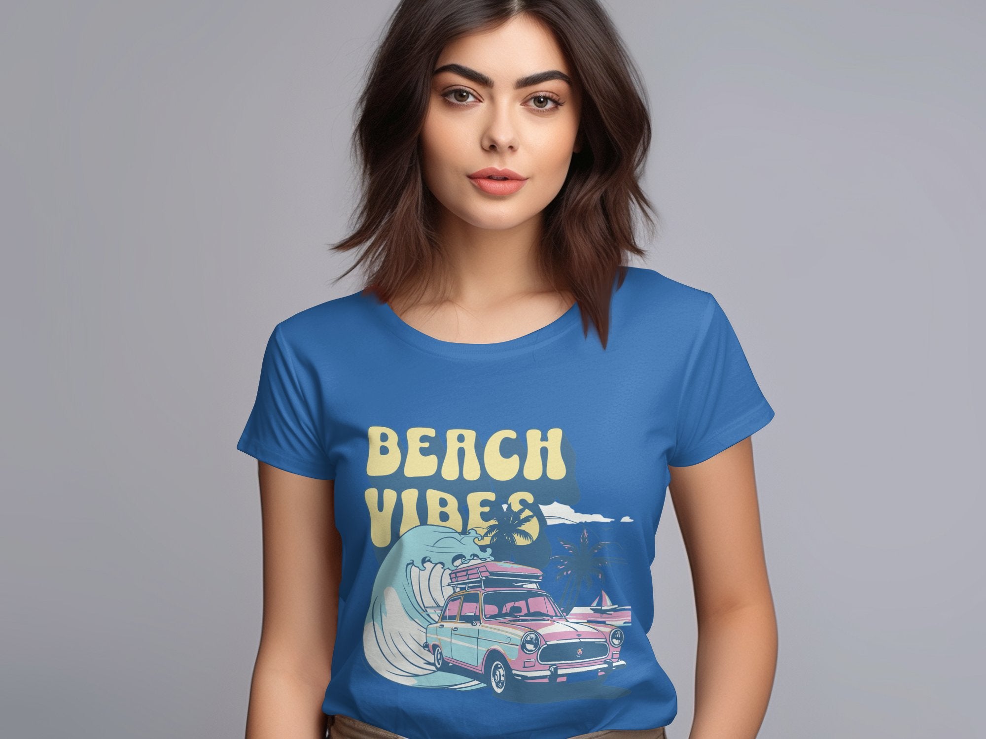 A woman with shoulder-length brown hair is wearing a blue Garment Graphics t-shirt featuring a retro surf design with a surfboard, car, waves, palm trees, and the text Beach Vibes. The background is plain and neutral.