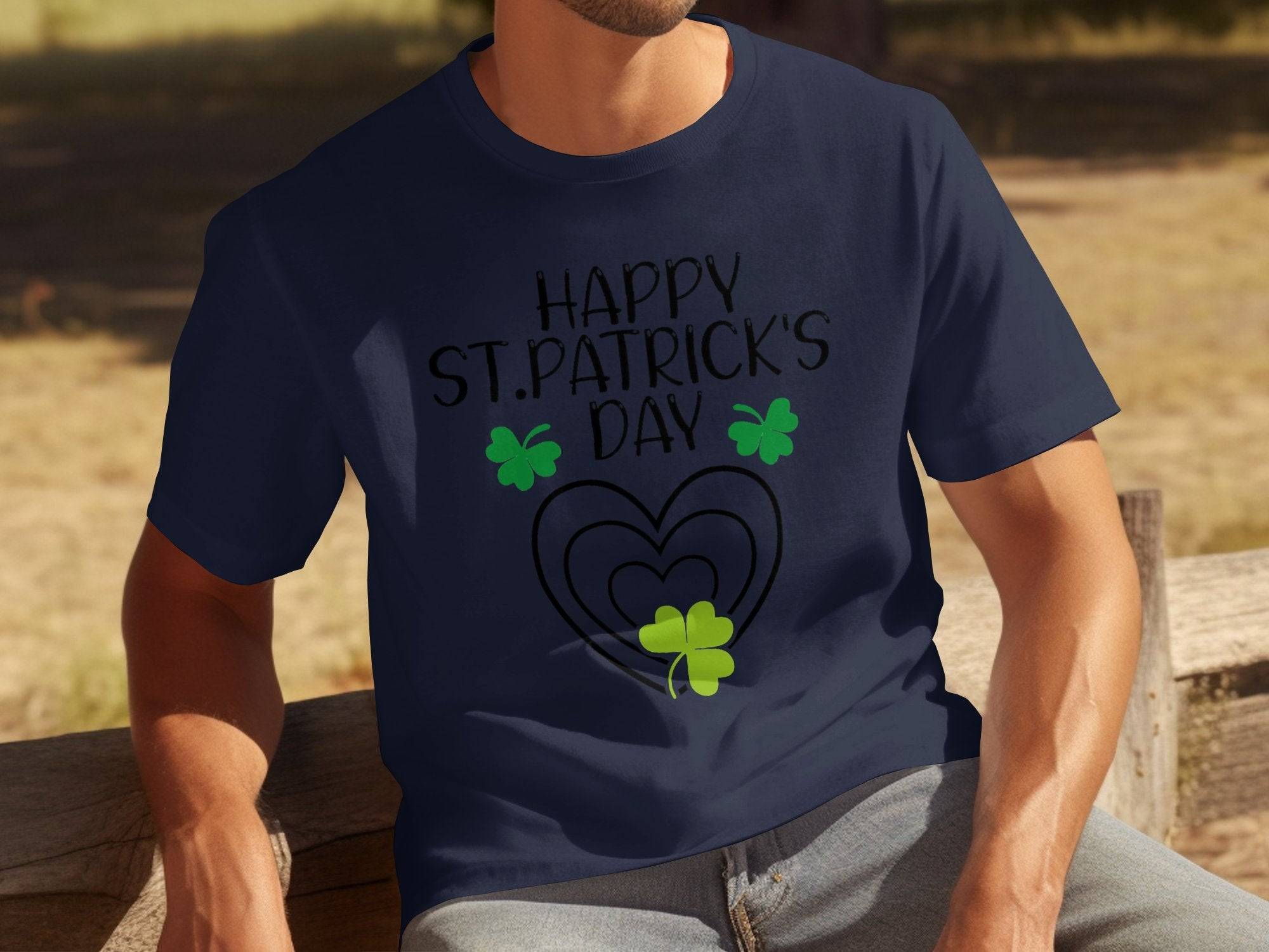 A man sits on a wooden bench outdoors, wearing a navy blue Garment Graphics t-shirt with Happy St. Patricks Day above three green shamrocks and a heart, adding festive flair to the grassy backdrop.