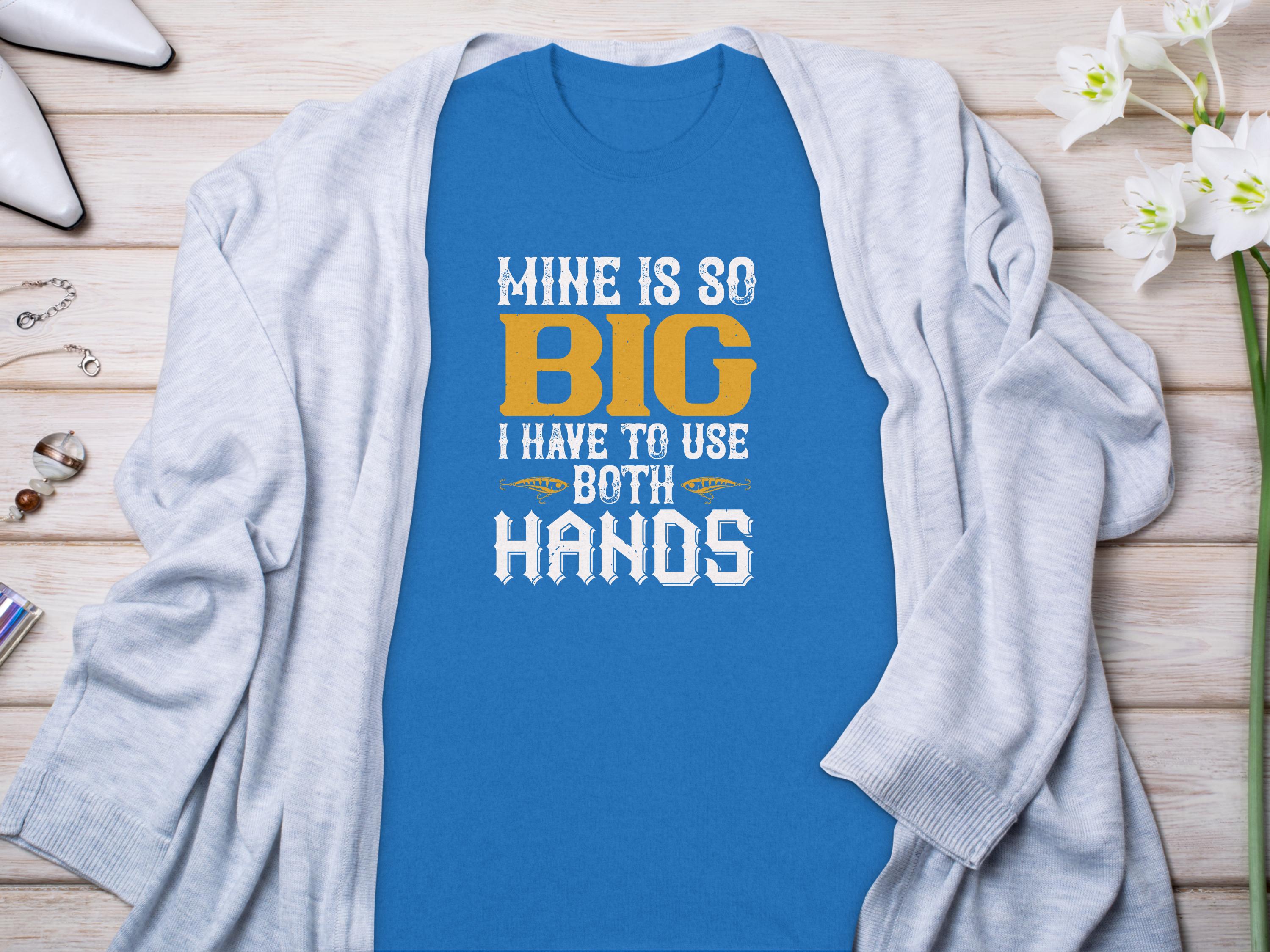 A classic fit blue graphic tee by Garment Graphics with white and gold text reading MINE IS SO BIG I HAVE TO USE BOTH HANDS sits on a wooden surface. Its paired with a light gray cardigan, white flowers, rings, a pen, and a wallet to highlight its humorous design.
.