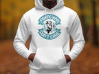 A person wears a Garment Graphics white hoodie featuring an anglers catch design and Fishing Hair Dont Care in teal, set against a blurred outdoor backdrop with trees and leaves.