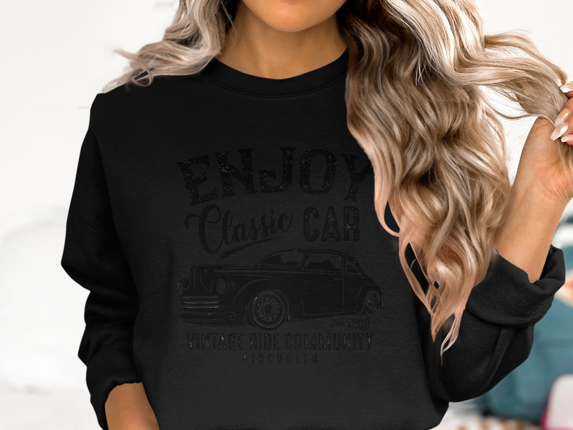 Sporting long wavy hair, a person wears a black sweatshirt from Garment Graphics featuring a classic car design with the words ENJOY Classic Car and Vintage Ride Community. The blurred white backdrop enhances the timeless charm of vintage rides.
