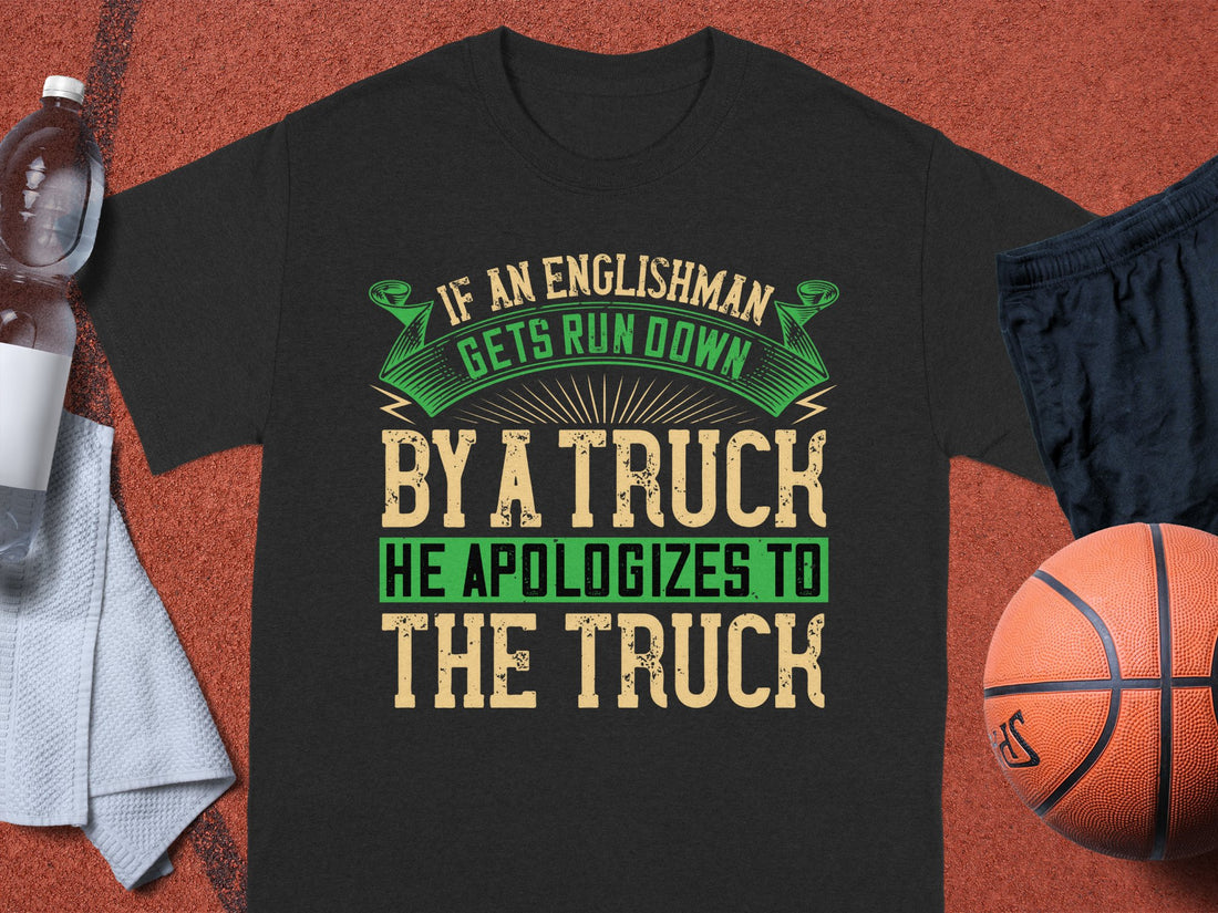 A Garment Graphics black t-shirt with a witty design: If an Englishman gets run down by a truck, he apologizes to the truck, is showcased next to a water bottle, cloth towel, basketball, and black athletic shorts.