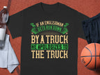 A Garment Graphics black t-shirt with a witty design: If an Englishman gets run down by a truck, he apologizes to the truck, is showcased next to a water bottle, cloth towel, basketball, and black athletic shorts.