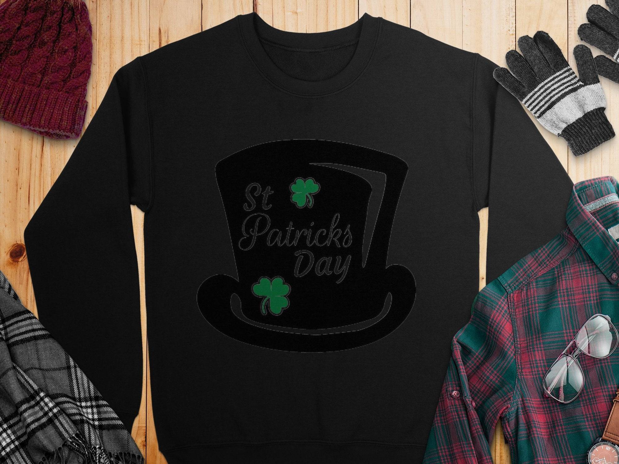 A Garment Graphics St. Patricks Day black sweatshirt with a top hat and shamrocks sits on wood, surrounded by a red knit hat, gray gloves, a black and white scarf, and a green and red plaid shirt, capturing the festive spirit.