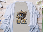 The Garment Graphics beige graphic T-shirt has a classic fit and features an ornate eye gaze design surrounded by gemstones and GAZE in bold. It rests on a light wooden surface, paired with a light gray cardigan adorned with white flowers and nearby accessories.