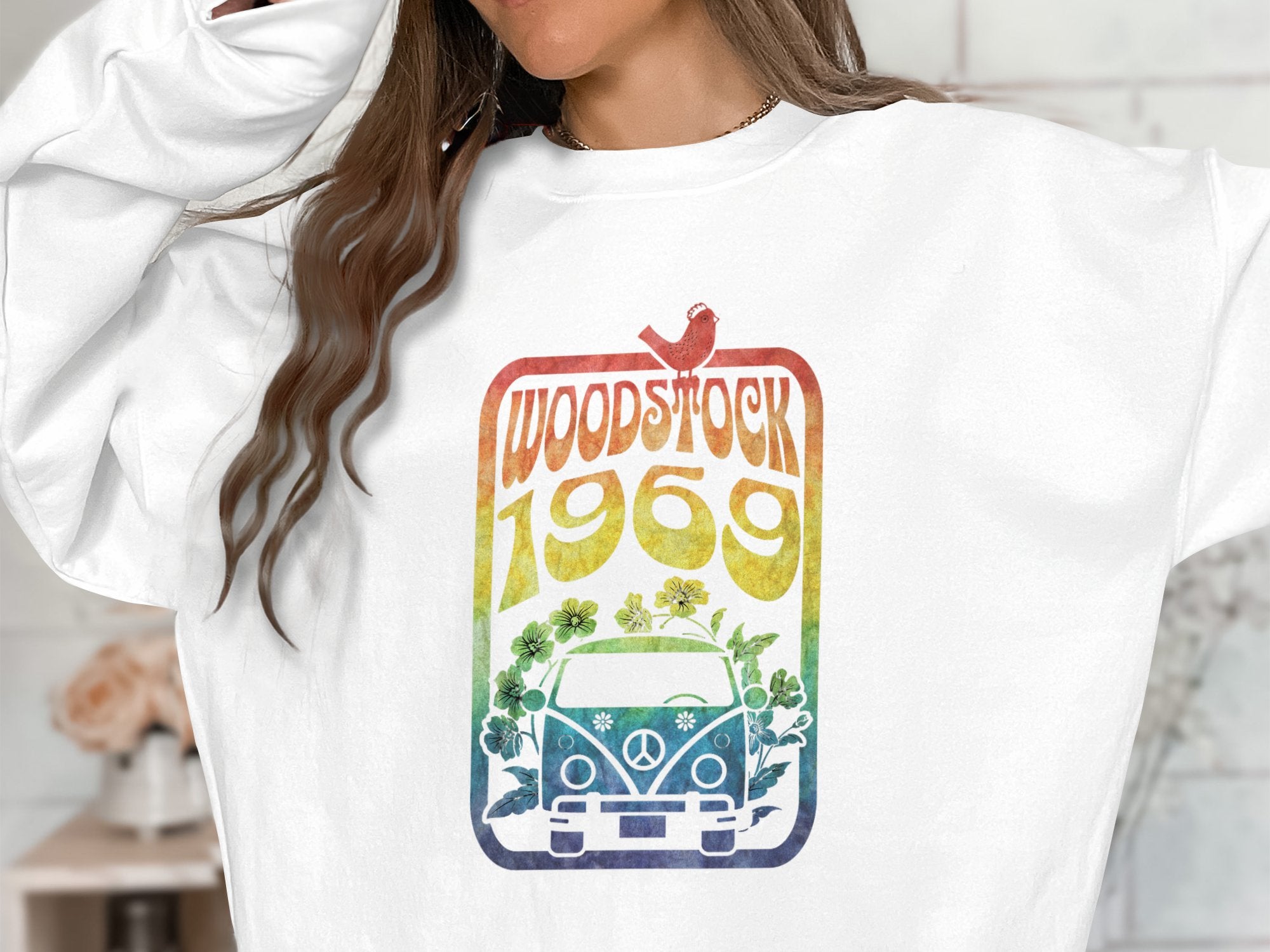 A person wears a Garment Graphics white sweatshirt featuring a vibrant retro design with WOODSTOCK 1969 above a flower-decorated van. A red bird sits on the text, adding charm to this vintage 1969 tribute.