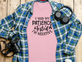 On a wooden surface lies a humorous Garment Graphics T-shirt with the text I Had My Patience Tested, Im Negative, exuding sarcasm. Its complemented by a blue and green plaid shirt, headphones, a camera, and a white coffee cup.