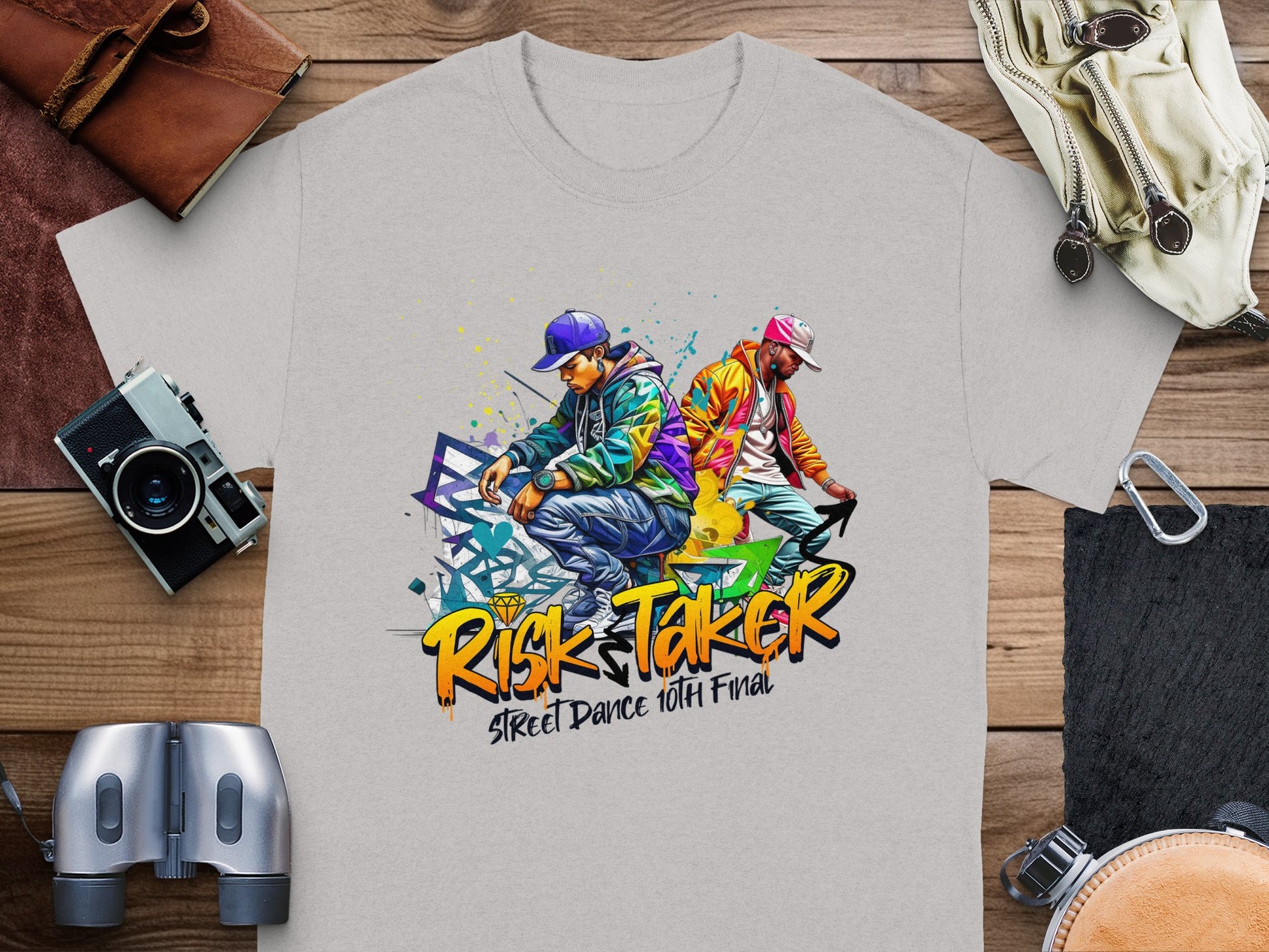 The Garment Graphics t-shirt features artistic designs of two street dancers in vibrant attire: one in purple and blue, the other in orange and green. The text Risk Taker Street Dance 10th Final highlights the essence of street dance culture, laid on a wooden surface.