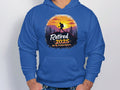 A person is wearing a blue hiking hoodie from Garment Graphics featuring a sunset mountain scene with trees, the words Retired 2025, Not My Problem Anymore, and a hikers silhouette.