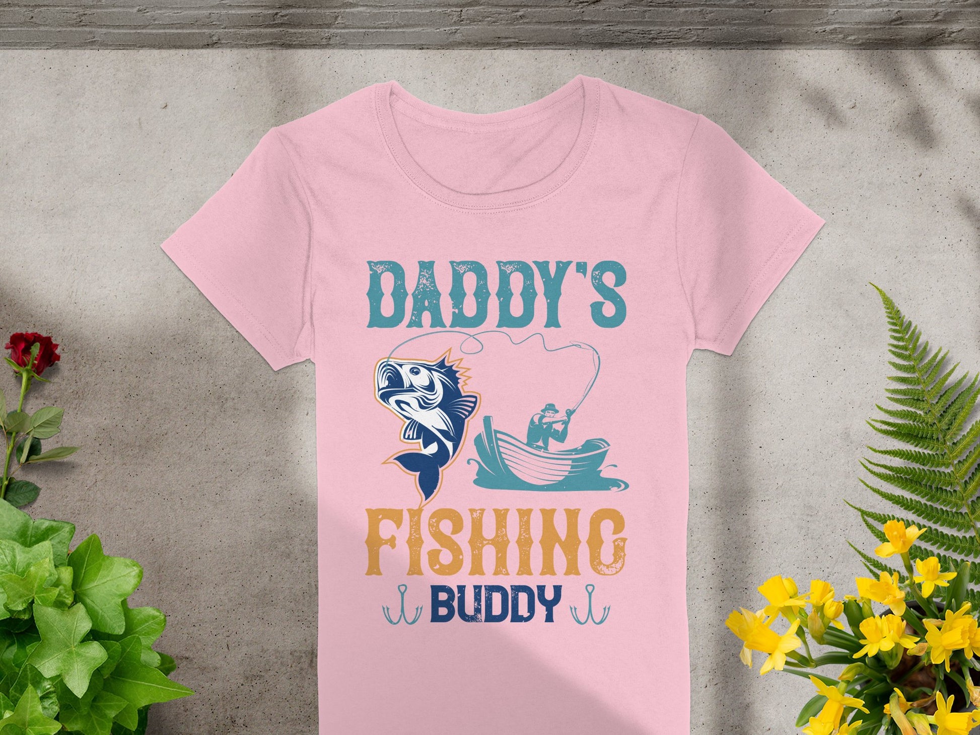 A pink womens fishing shirt by Garment Graphics features Daddys Fishing Buddy with a playful graphic of a fish and person casting from a boat, set against concrete with vibrant greenery and yellow flowers.