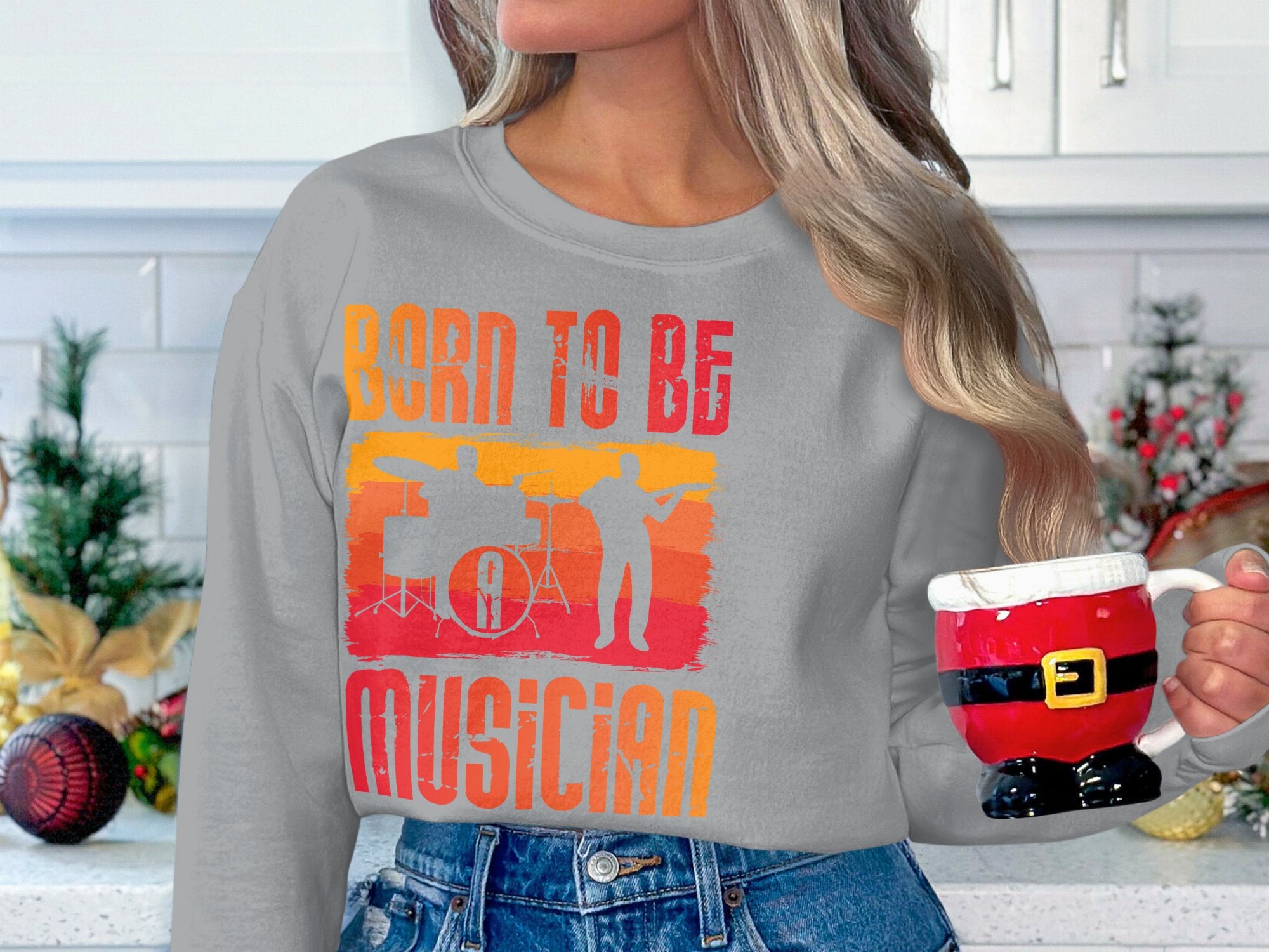 Clad in a Garment Graphics gray sweatshirt with Born to Be Musician, a person holds a Santa-themed mug amid festive decor, including pine branches and ornaments. Perfect for music lovers.