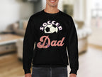 A person is wearing a stylish Soccer Dad sweatshirt by Garment Graphics, featuring unisex sizing and a soccer ball image against a blurred indoor backdrop with furniture.