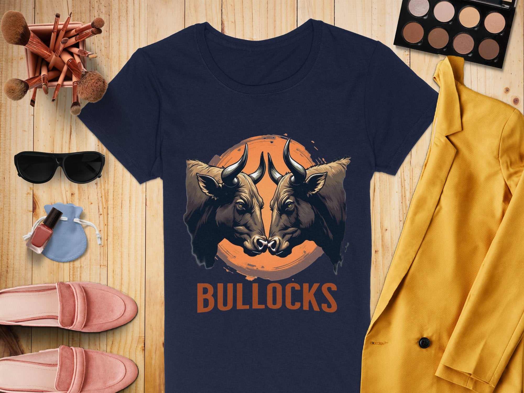 A navy T-shirt from Garment Graphics, adorned with bold bull illustrations and BULLOCKS, is laid out on a wooden surface. Its paired with makeup brushes, eyeshadow, sunglasses, a yellow jacket, pink shoes, and nail polish—ideal for creating a casual wear vibe.