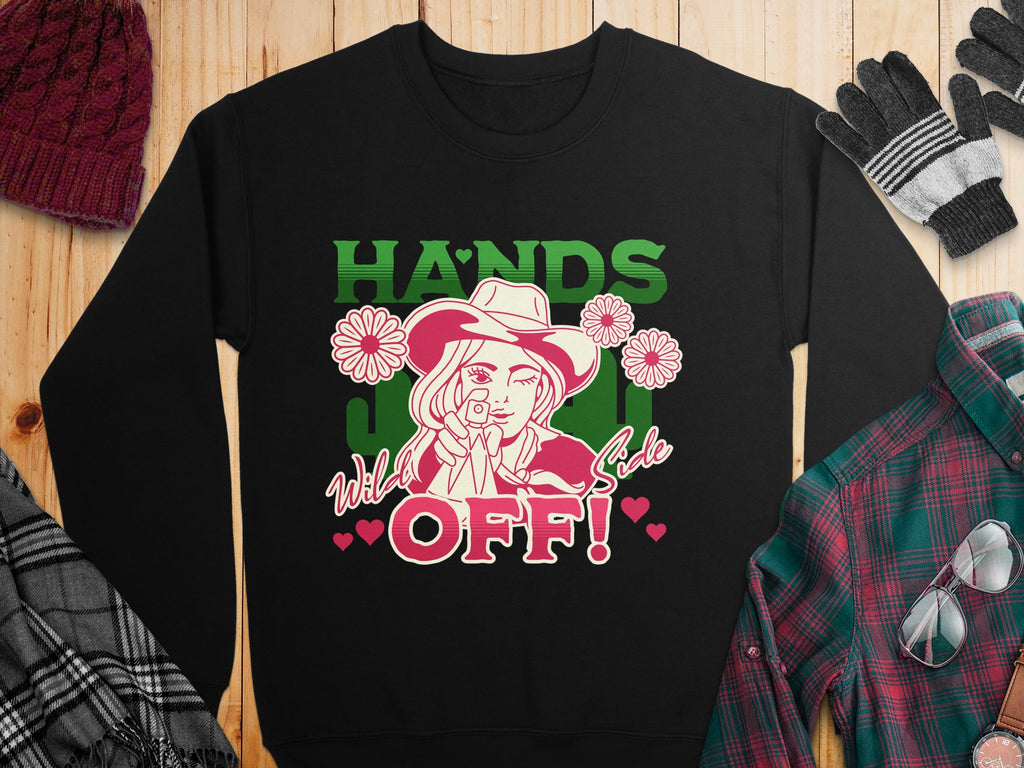 Garment Graphics offers a black loose-fit sweatshirt featuring a line drawing of a person in a hat with flowers and a lasso. The phrase HANDS OFF! Wild Side appears in green, pink, and white. Nearby are accessories: red knit hat, plaid shirt, black gloves, and gray scarf.