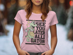 A woman wearing a light pink Garment Graphics T-shirt featuring a cartoon alligator and the playful phrase See You Later Alligator stands against an artfully blurred backdrop of warm and cool tones.